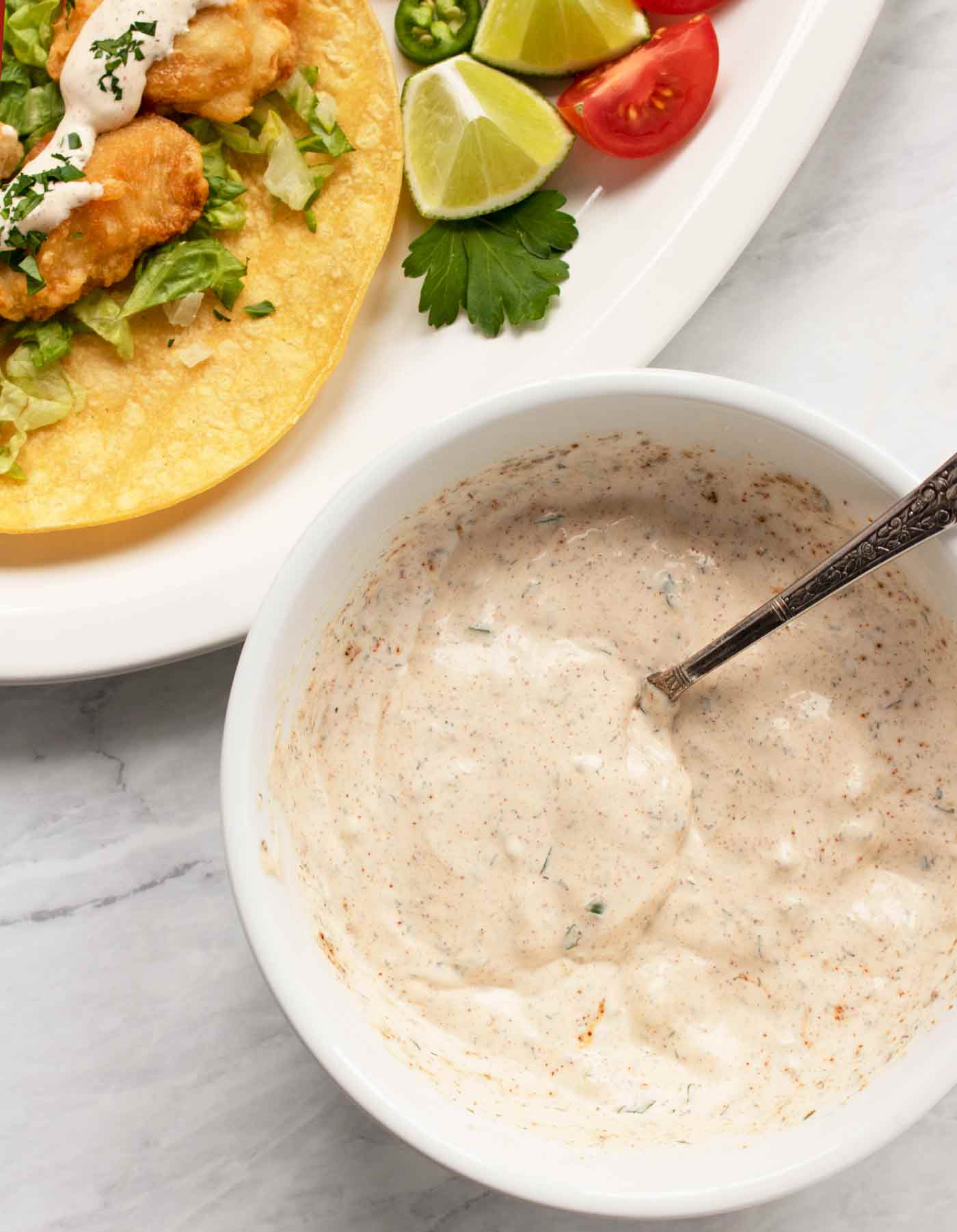 A bowl of fish taco sauce with a spoon next to a plate of fish tacos.