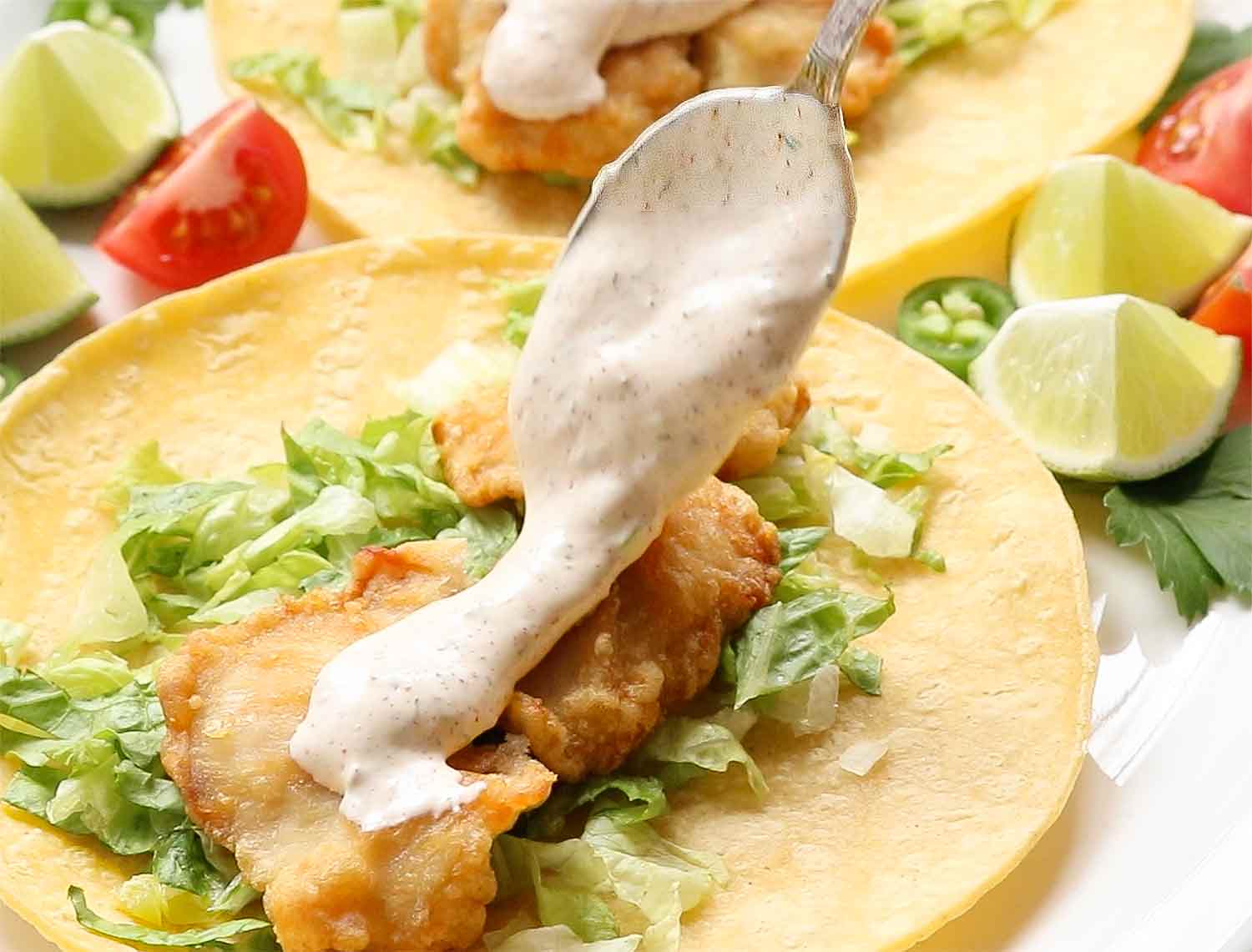 Spooning fish taco sauce over a taco with crispy fried fish and lettuce.
