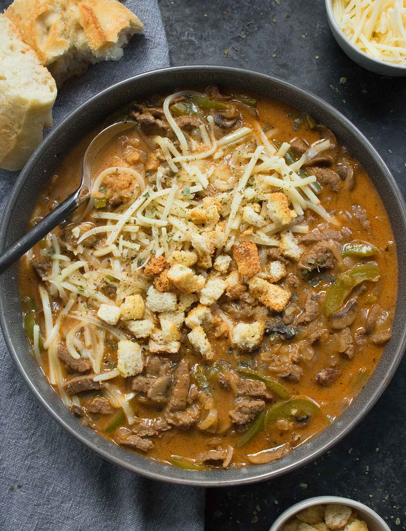 Philly Cheesesteak Soup in a gray bowl with toppings on the side.