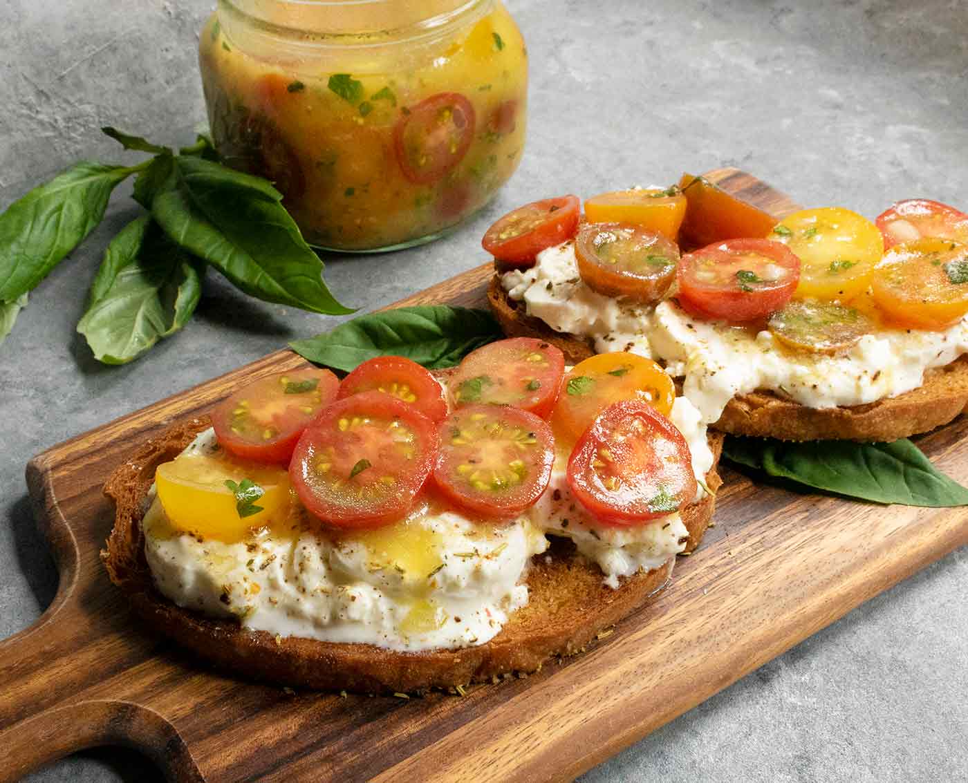 Side view of two slices of cherry tomato bruschetta, with a jar of marinated tomatoes in the background
