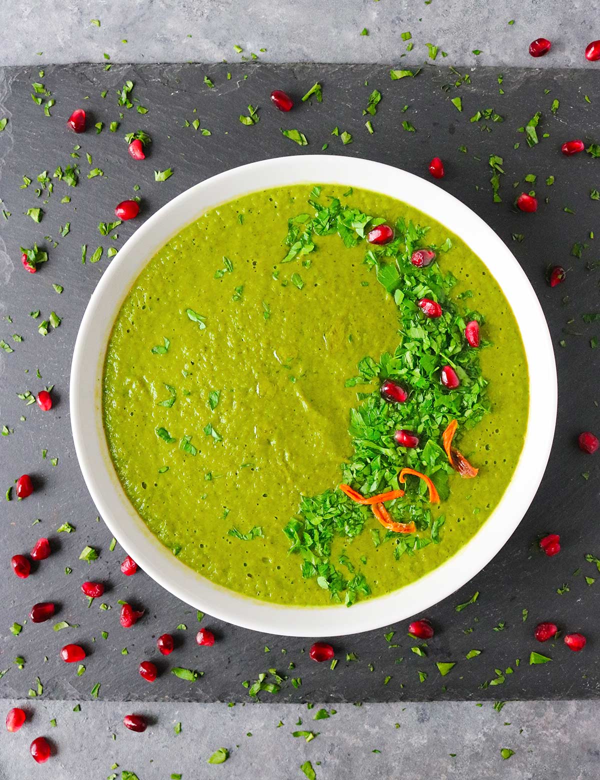 Overhead view of Green Soup in a white bowl, topped with herbs and pomegranate seeds.