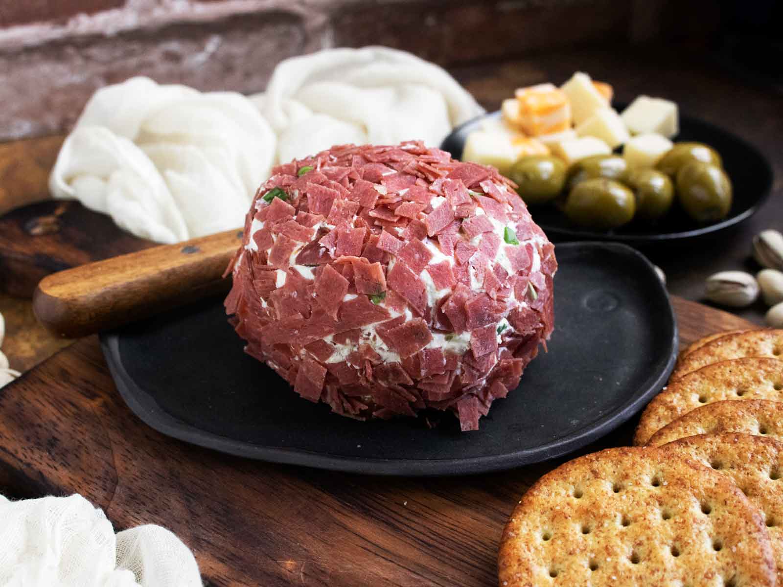A Dried Beef Cheese Ball on a small black appetizer plate with a spreading knife and crackers, and cheeses and olives in the background.