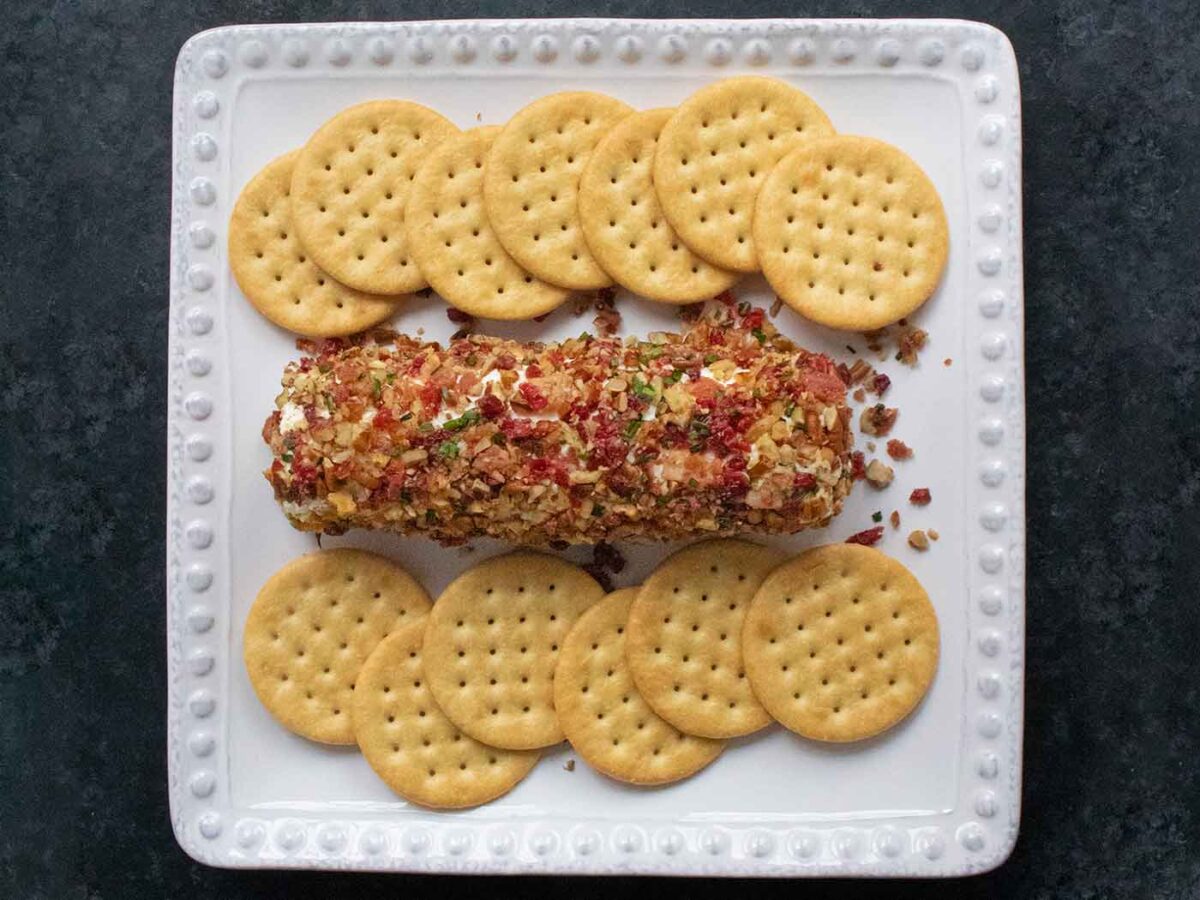 Overhead view of Cranberry Pecan Bacon Cheese Log on a square white plate with crackers.