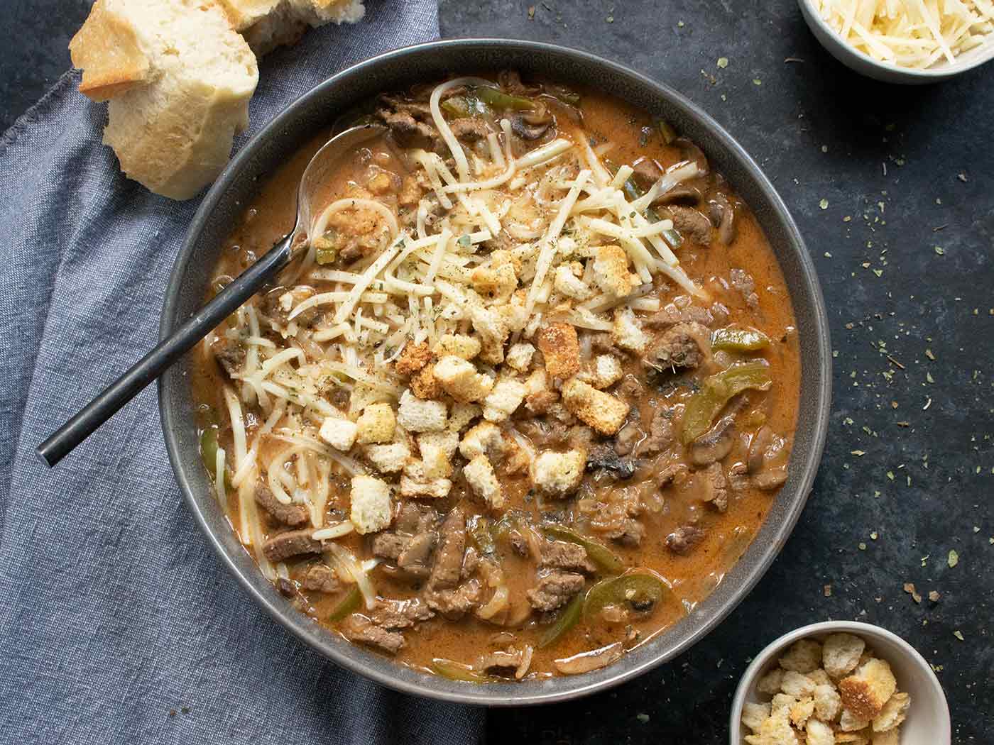 Overhead view of Philly Cheesesteak Soup in a dark gray bowl with a rustic spoon.