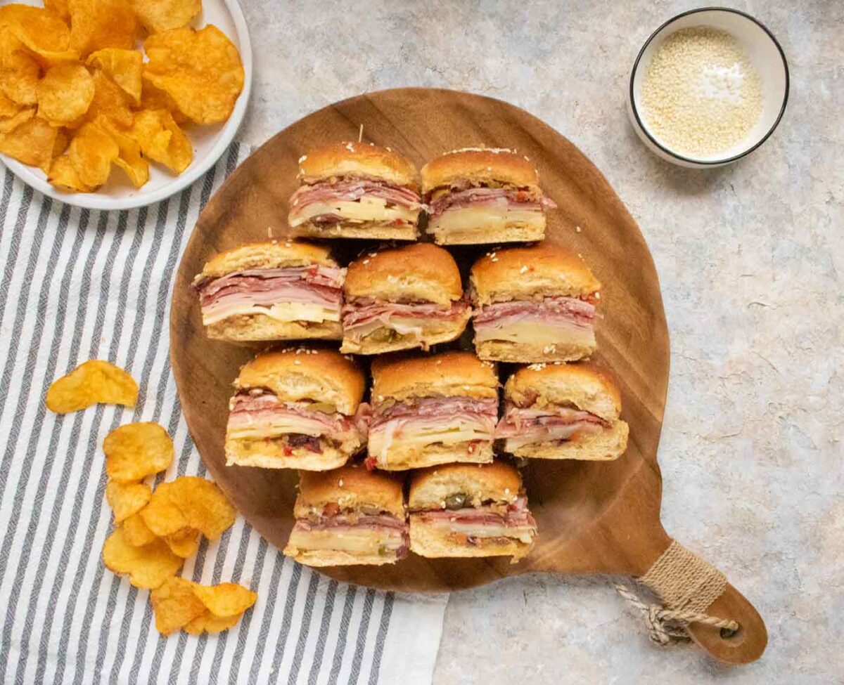 Muffuletta Sliders on a wooden serving board with potato chips on the side.