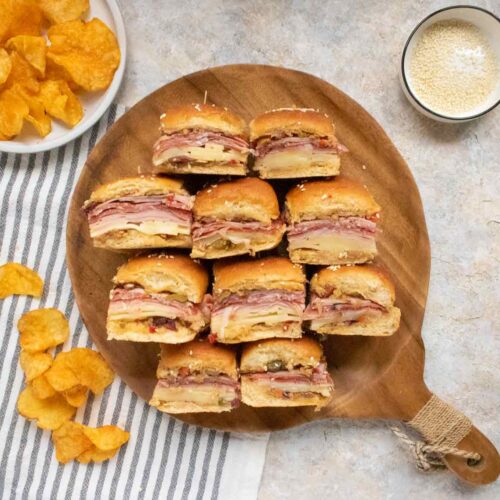 Muffuletta Sliders on a wooden serving board with potato chips on the side.