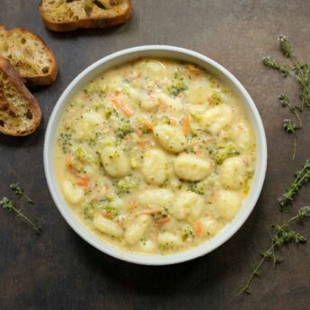 Overhead view of Broccoli Cheddar Gnocchi Soup in a white bowl with a side of sliced bread.