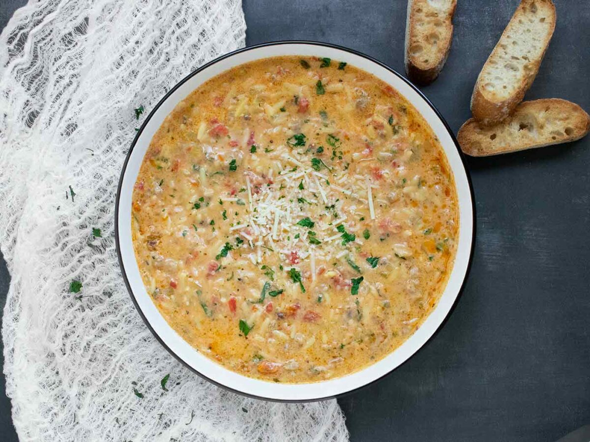 An overhead view of a Marry Me Sausage Orzo Soup in a white bowl with a black rim, and baguette slices on the side.