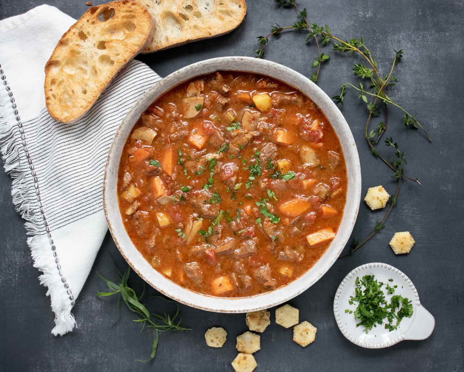 Overhead view of Braised Vegetable Beef Soup in a rustic gray bowl with bread slices on the side.