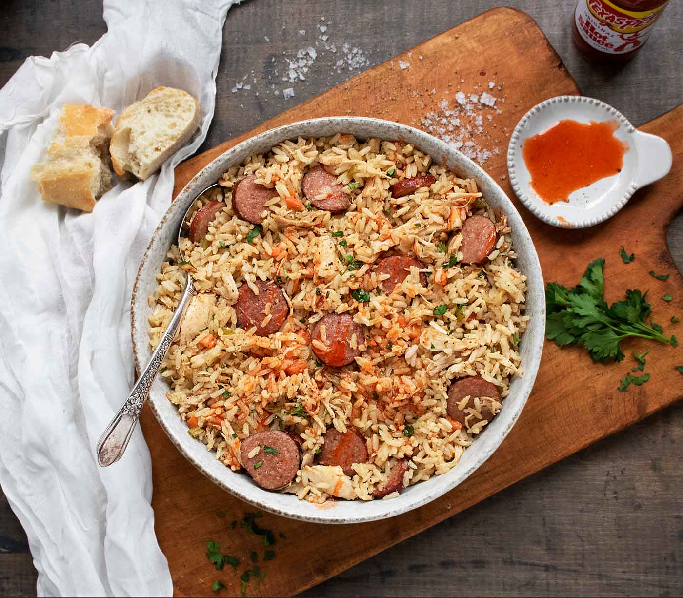 An overhead view of Chicken Bog in a bowl, with hot sauce and a sliced baguette served on the side.