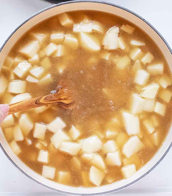 Potatoes and broth added to the pot, stirring with a wooden spoon.