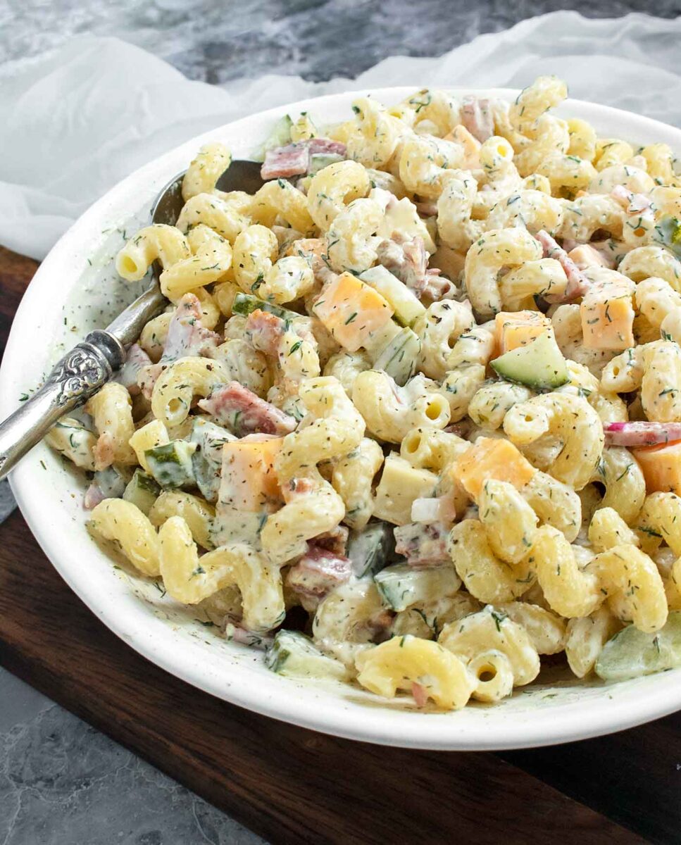 Side-angled view of Dill Pickle Pasta Salad in a white bowl with a serving spoon.