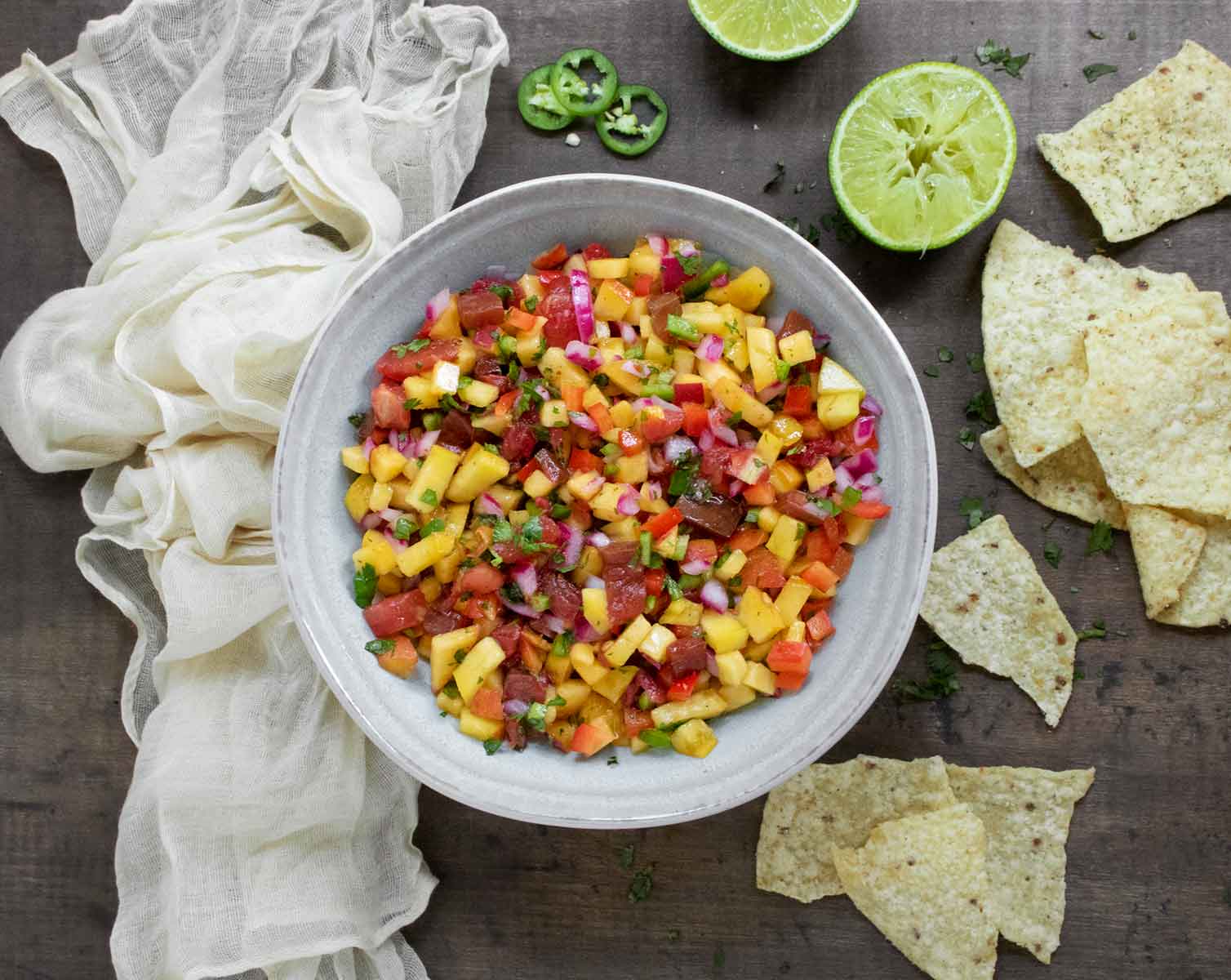 An overhead view of Peach Salsa in a pale blue-gray bowl with tortilla chips and lime halves on the side.