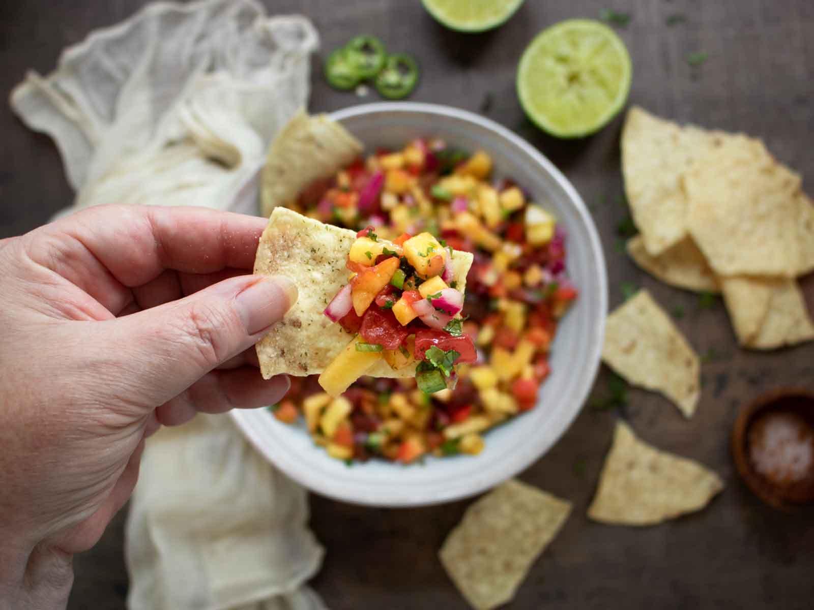 Hand holding a tortilla chip loaded with peach salsa, with the bowl of salsa in the background.