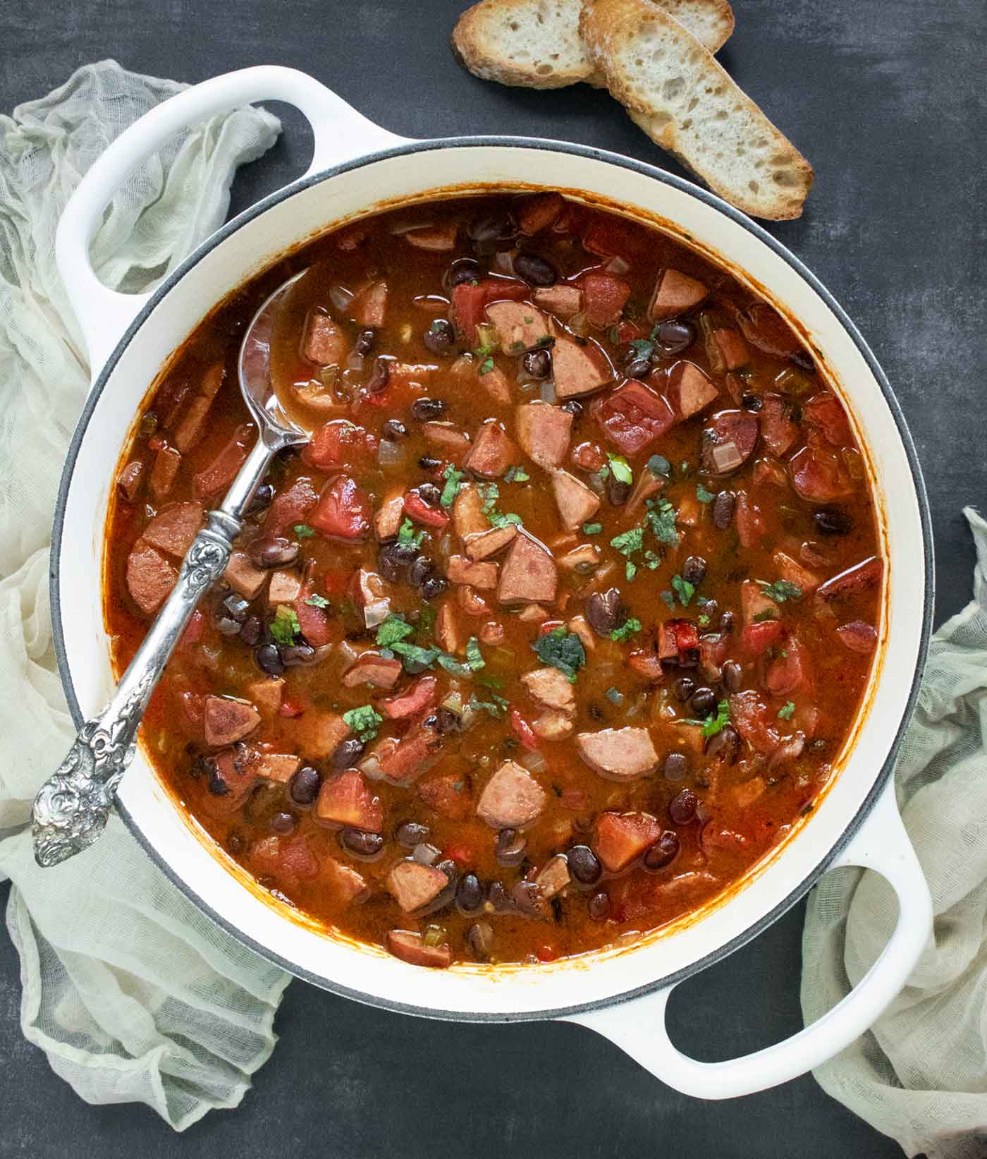 Overhead view of a pot of Smoky Black Bean and Sausage Soup with a serving spoon, and a side of sliced bread.