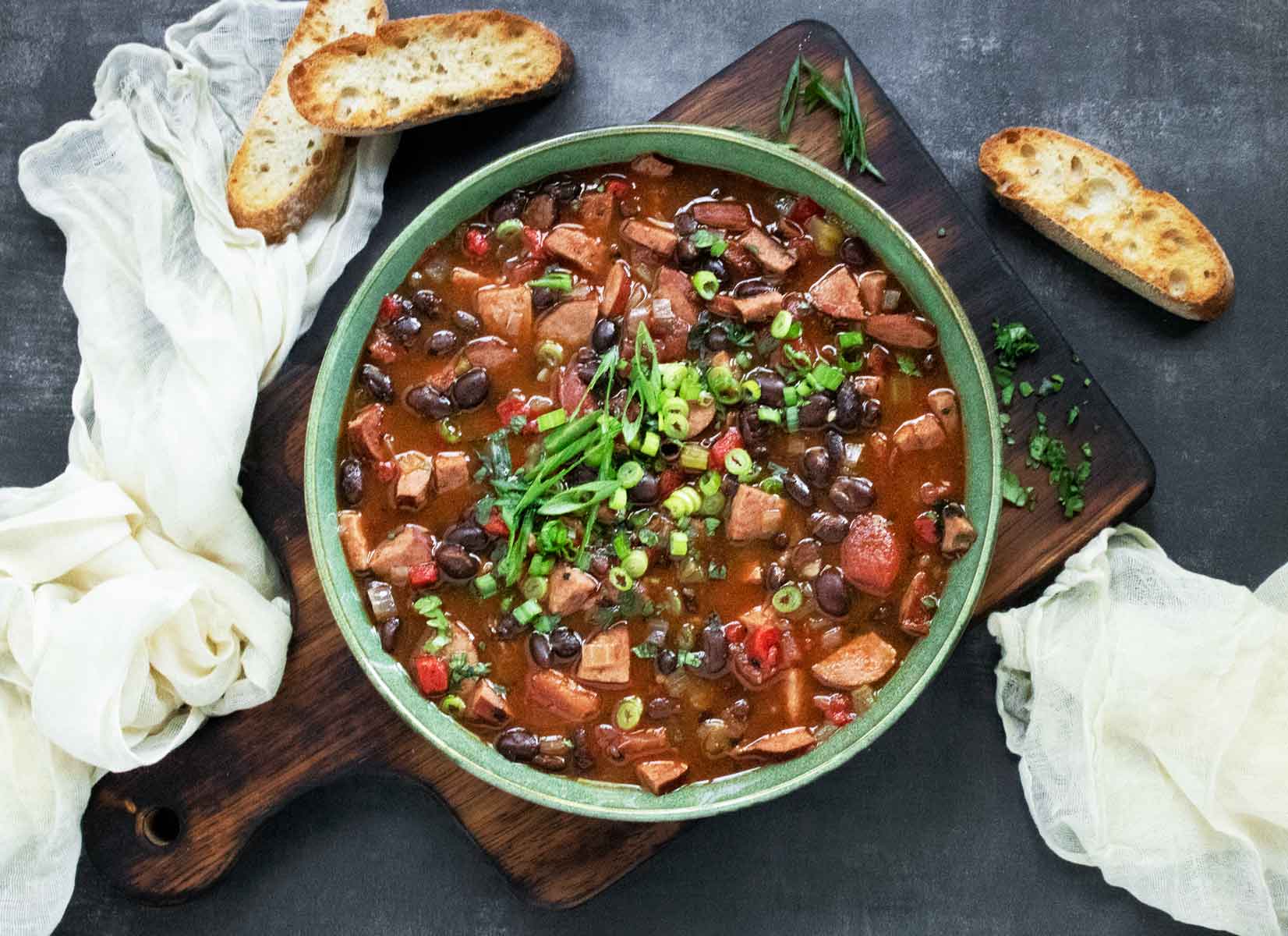 Overhead view of Smoky Black Bean and Sausage Soup in a green bowl on a wooden board.