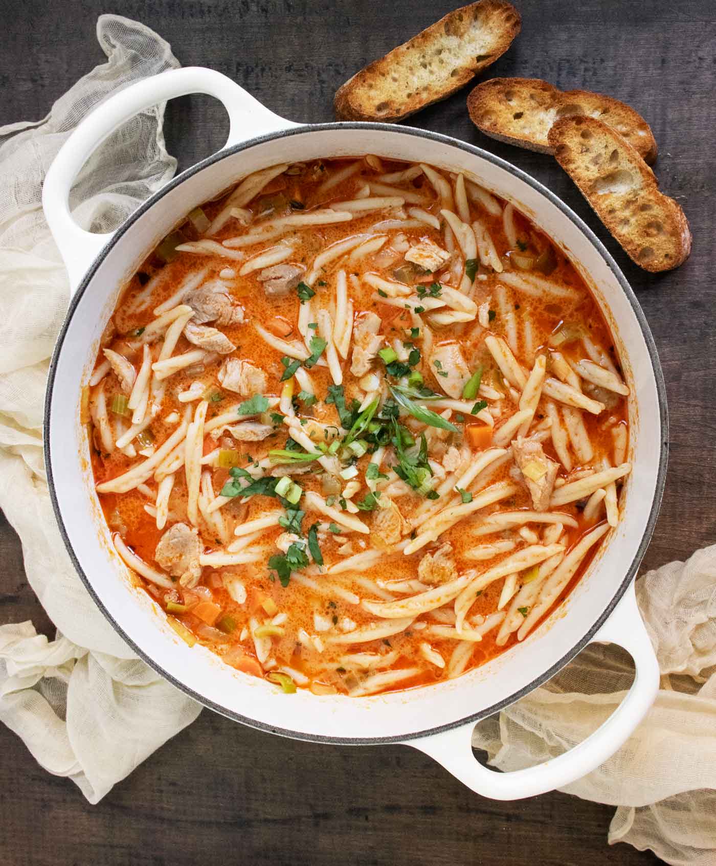 An overhead view of Thai Fusion Chicken Noodle Soup in a large white Dutch oven with slices of bread on the side.