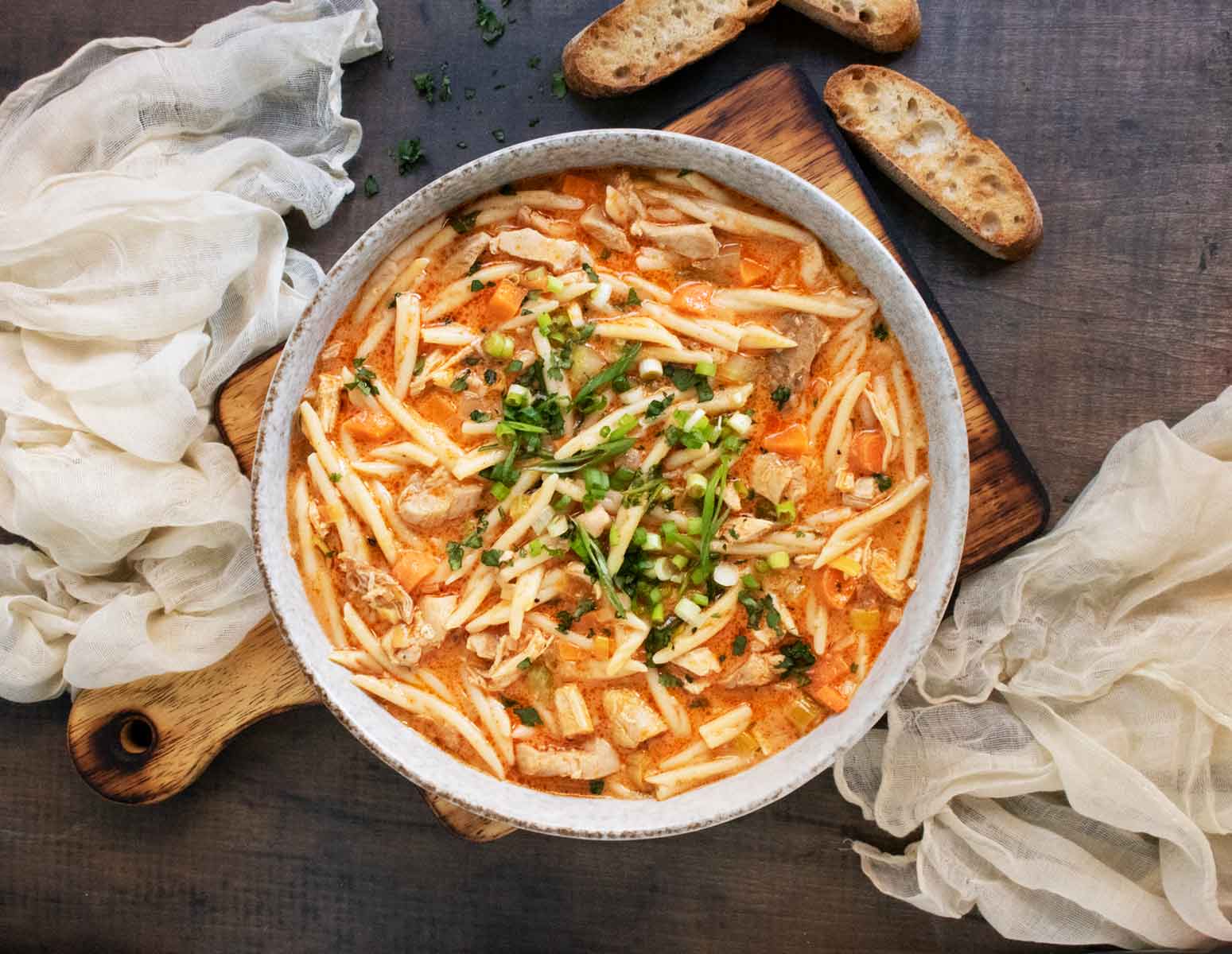 Overhead view of a bowl of Thai Fusion Chicken Noodle Soup on a wooden board with a side of bread slices.