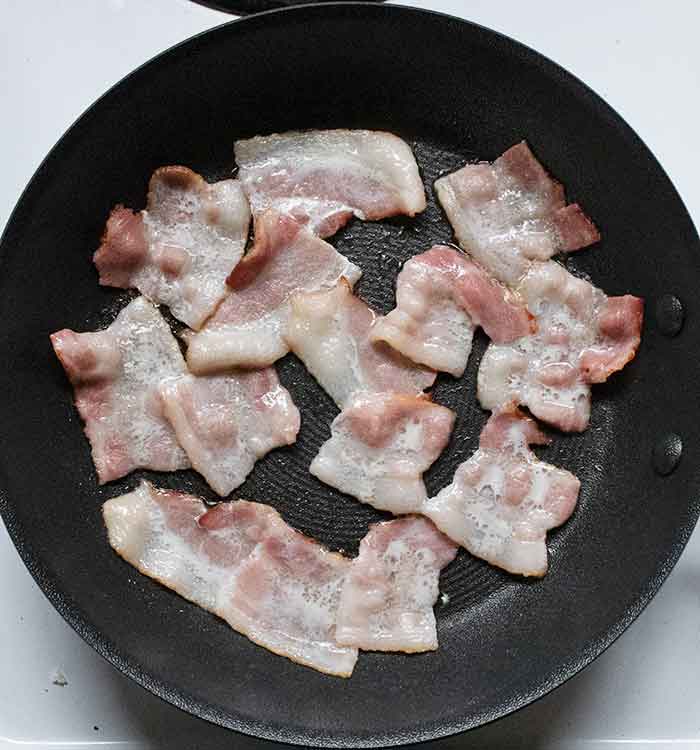 Short pieces of raw bacon arranged in a skillet, starting to sizzle.