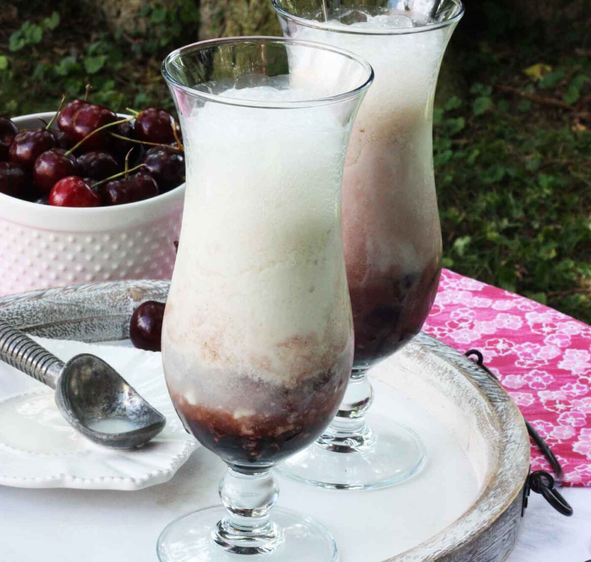Two tall glasses of Black Cherry Champagne Floats on a white serving board, taken outside.