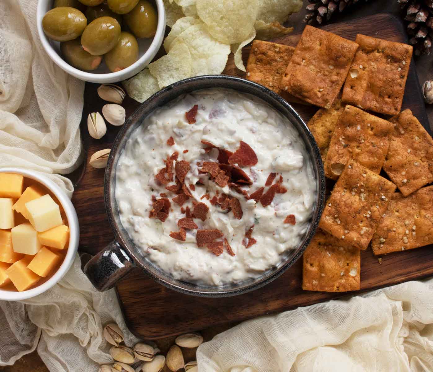 Overhead view of Crispy Pepperoni Ranch Dip in a black ceramic bowl, on a wooden serving board with a variety of chips, cheeses and olives.