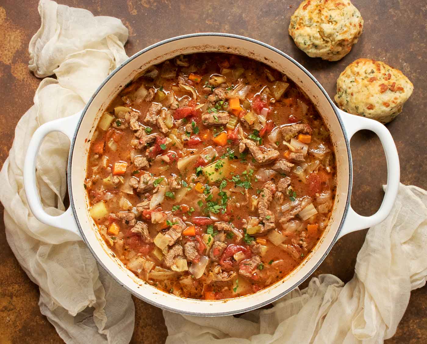 Overhead view of Beef and Cabbage Soup in a large, white Dutch oven, with a side of cheddar biscuits.