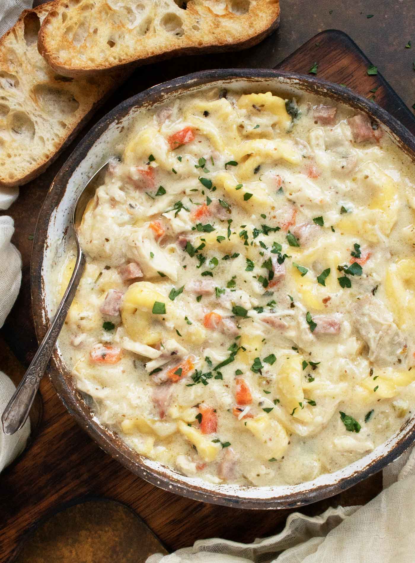 Close-up of a bowl of Cordon Bleu Tortellini Soup with a spoon, and two crostini slices on the side.