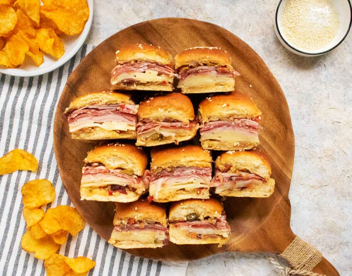 Muffuletta Sliders turned on their sides so you can see the fillings, arranged on a round wooden serving board.