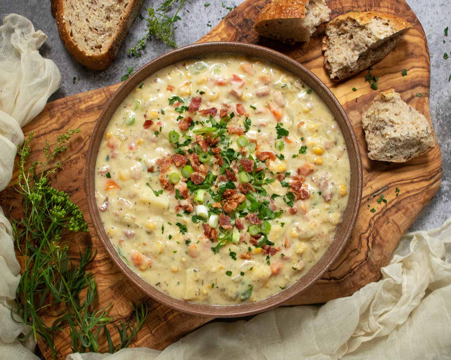 Overhead view of Ham and Corn Chowder in a brick-red bowl on a wooden serving board, with chunks of bread on the side.