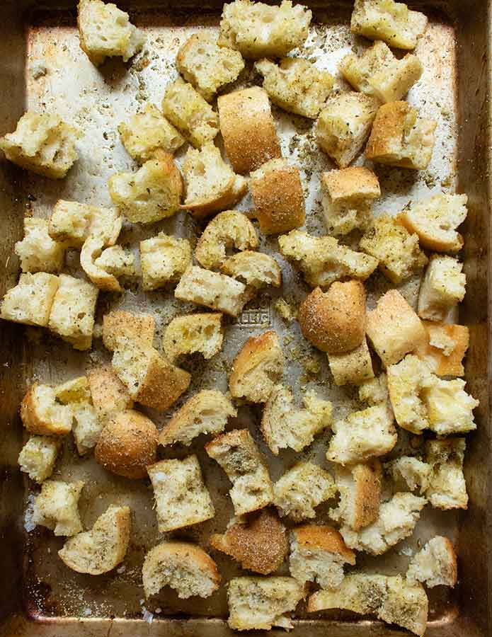 Seasoned bread pieces spread out on a baking sheet, ready to go into the oven.