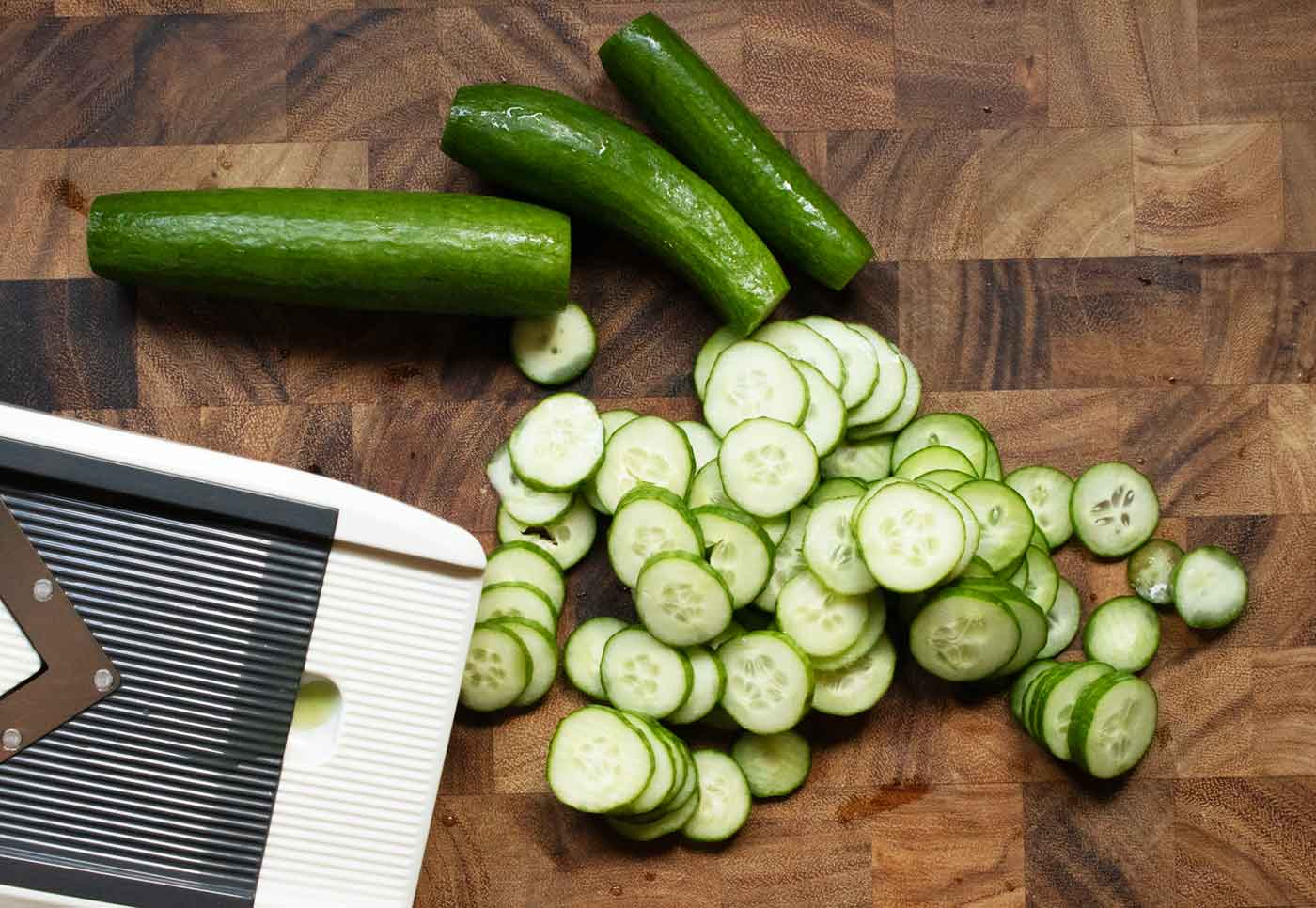 Cucumber slices and whole cucumbers on a wooden cutting board, with a mandoline.