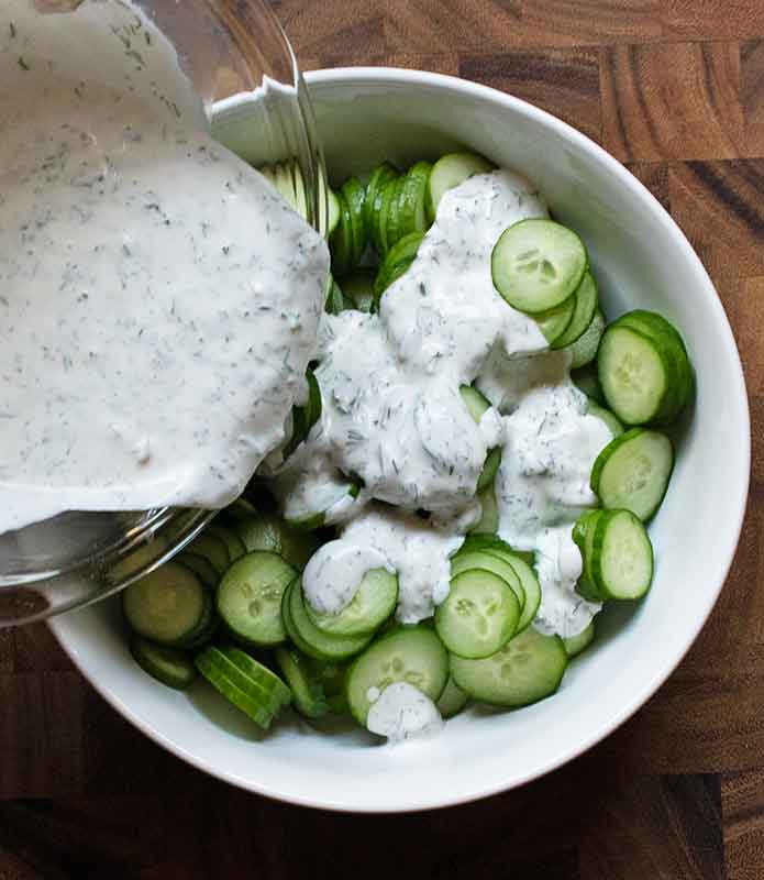 Pouring the tzatziki dressing over the cucumber slices in a large white bowl.