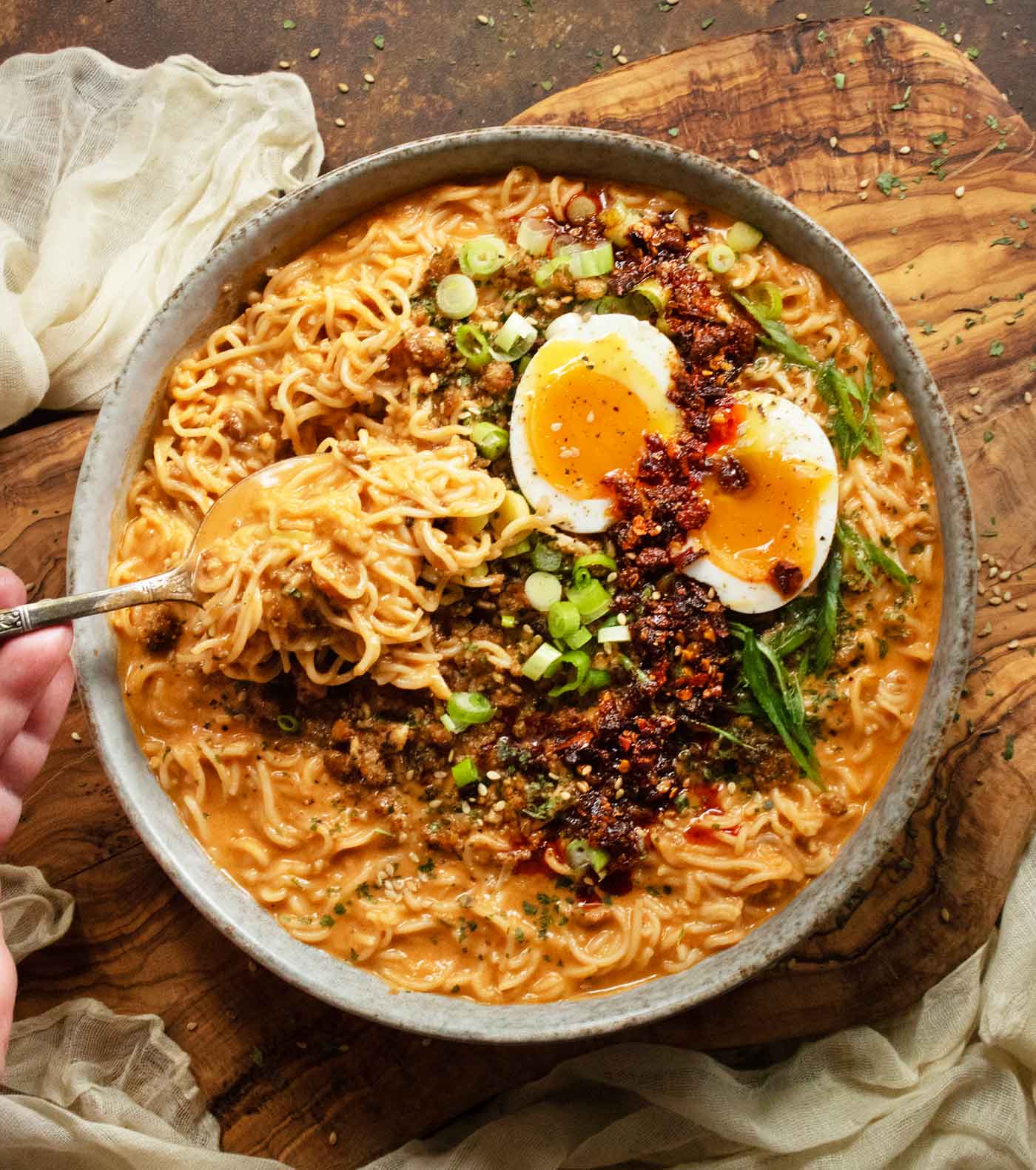 Overhead view of Spicy Peanut Chili Crisp Ramen Noodle Soup in a rustic bowl on a wooden board. My hand is holding a spoon loaded with ramen noodles.