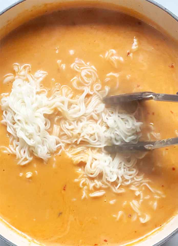 Adding the cooked ramen to the soup using tongs.